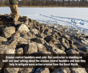a person standing on a pile of rock in the snow. It has the text, "Erosion control boulders west side: Our contracter is standing on built-out land talking about the erosion control boulders and how they help to mitigate wave action erosion from the Great Marsh."