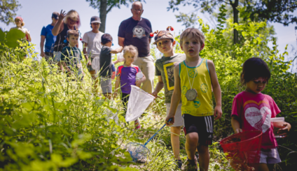 An image of a group of children wandering on a path through Fontenelle Forest with butterfly nets; one of them is wearing a hat with bug antennae on top.